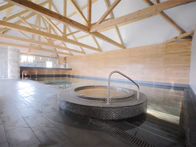 An indoor swimming pool with a built-in jacuzzi inside a large, purpose built room with large wooden beams.