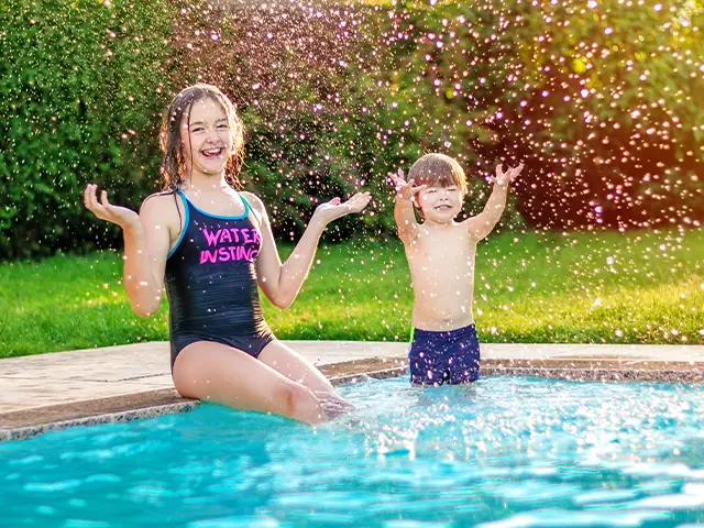 A brother and sister splashing in their garden swimming pool in the early evening light.