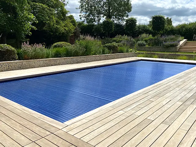 An outdoor swimming pool with the safety cover pulled across set in decking in a nicely kept garden.