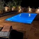 A garden swimming pool surrounded by a patio lit up at night.