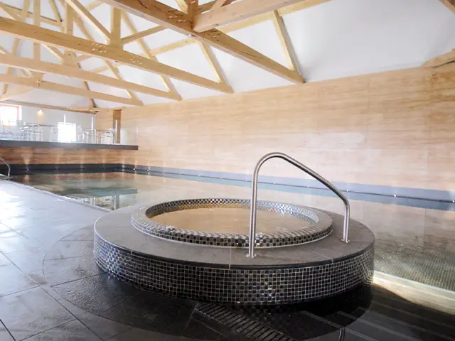An indoor swimming pool with a built-in jacuzzi inside a large, purpose built room with large wooden beams.